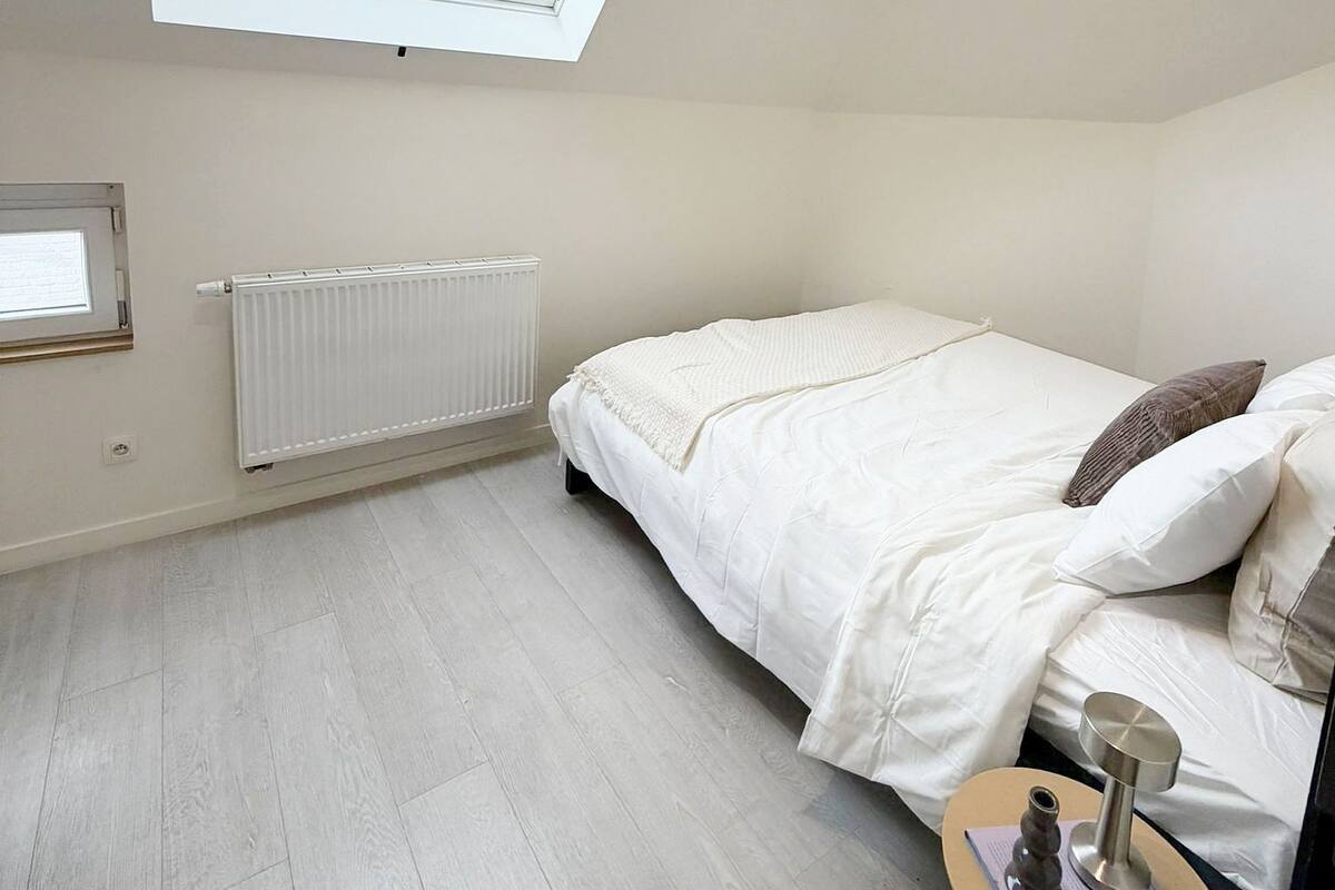 Een slaapkamer met dakraam, een radiator, een bed met kussens, een nachtkastje met vaas en uitzicht op gebouwen.