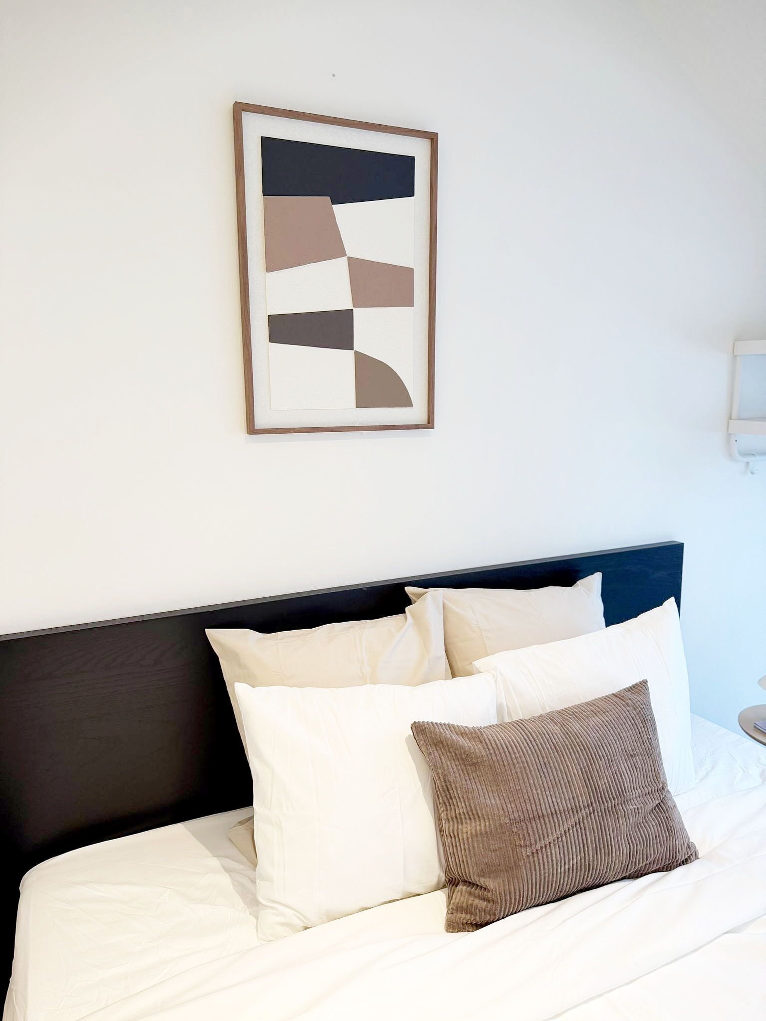 Una cama con ropa de cama blanca y un cabecero oscuro, una obra de arte abstracta enmarcada en la pared y una mesita de noche con una lámpara.