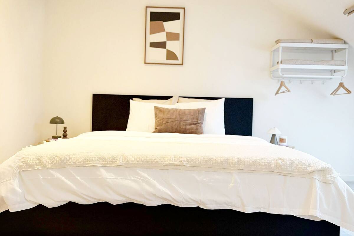Una cama con cabecera oscura, ropa de cama blanca, un solo almohadón y una mesita de noche con una lámpara.