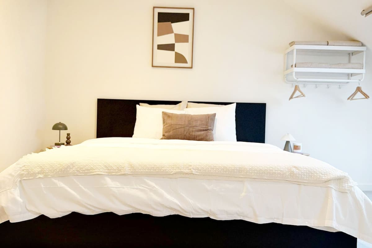 Een bed met een donker hoofdeinde, witte beddengoed, één kussen en een nachtkastje met een lamp.