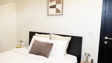 Een net opgemaakt bed met wit en bruin beddengoed, een modern zwart hoofdbord, een lijst met abstracte kunst aan de muur en een klein nachtkastje met een lamp en decoratieve objecten.