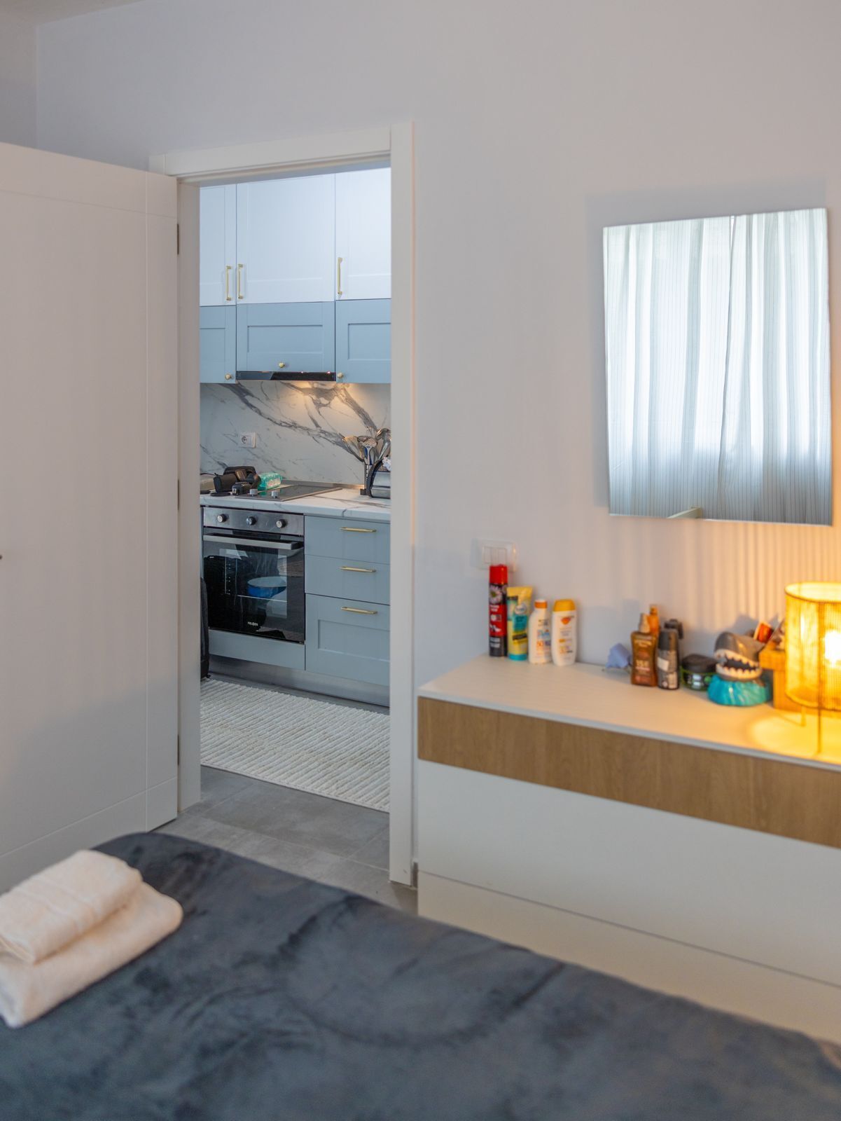 A bedroom with a bed, a bedside table with toiletries, a window with curtains, and a view into a kitchen with cabinets and a stove.
