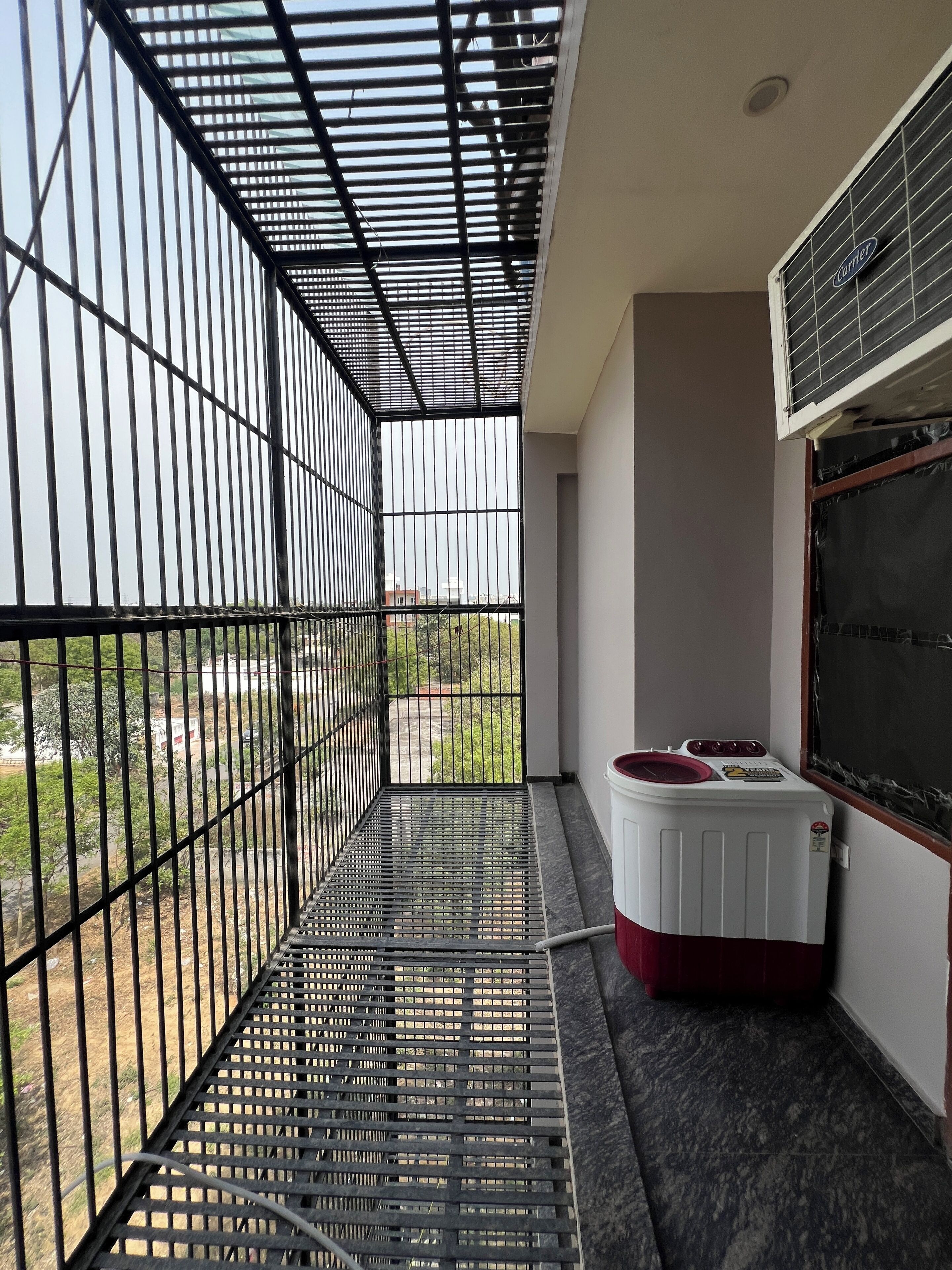 Een balkon met een metalen hekwerk, een wasmachine en een raam met een Carrier airconditioner.