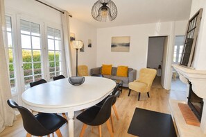 Ein Essbereich mit einem runden Tisch, schwarzen Stühlen und einer weißen Tischdecke. Im Hintergrund befindet sich eine gelb gepolsterte Couch und ein Gemälde an der Wand.
