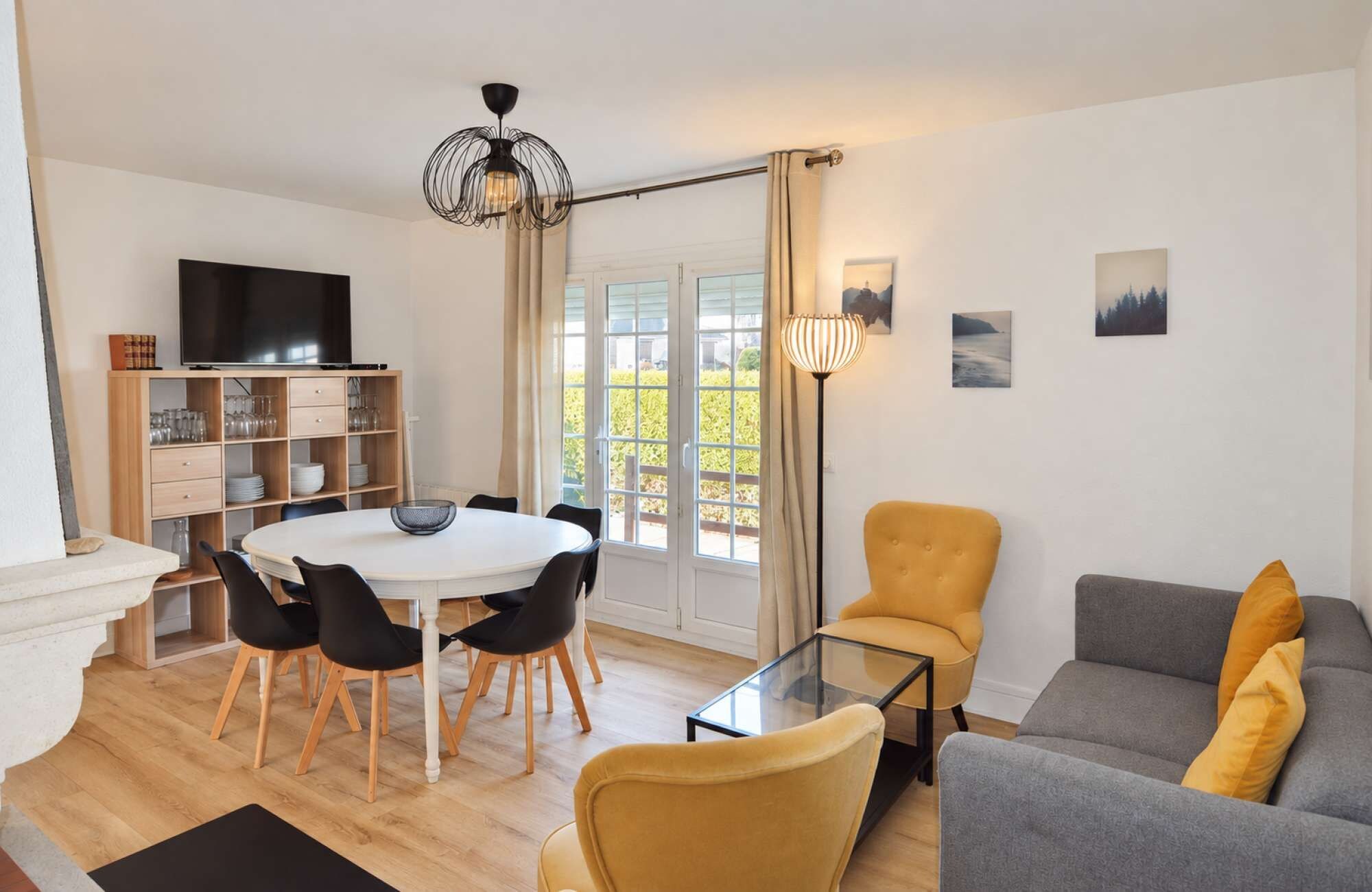 Un salon moderne avec une table à manger ronde, des chaises noires, un fauteuil jaune, un canapé gris et un téléviseur à écran plat posé sur une étagère en bois.