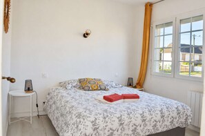 Ein Schlafzimmer mit einem Bett, einem Nachttisch, einer Lampe, einem Fenster mit Vorhängen und Blick ins Freie.