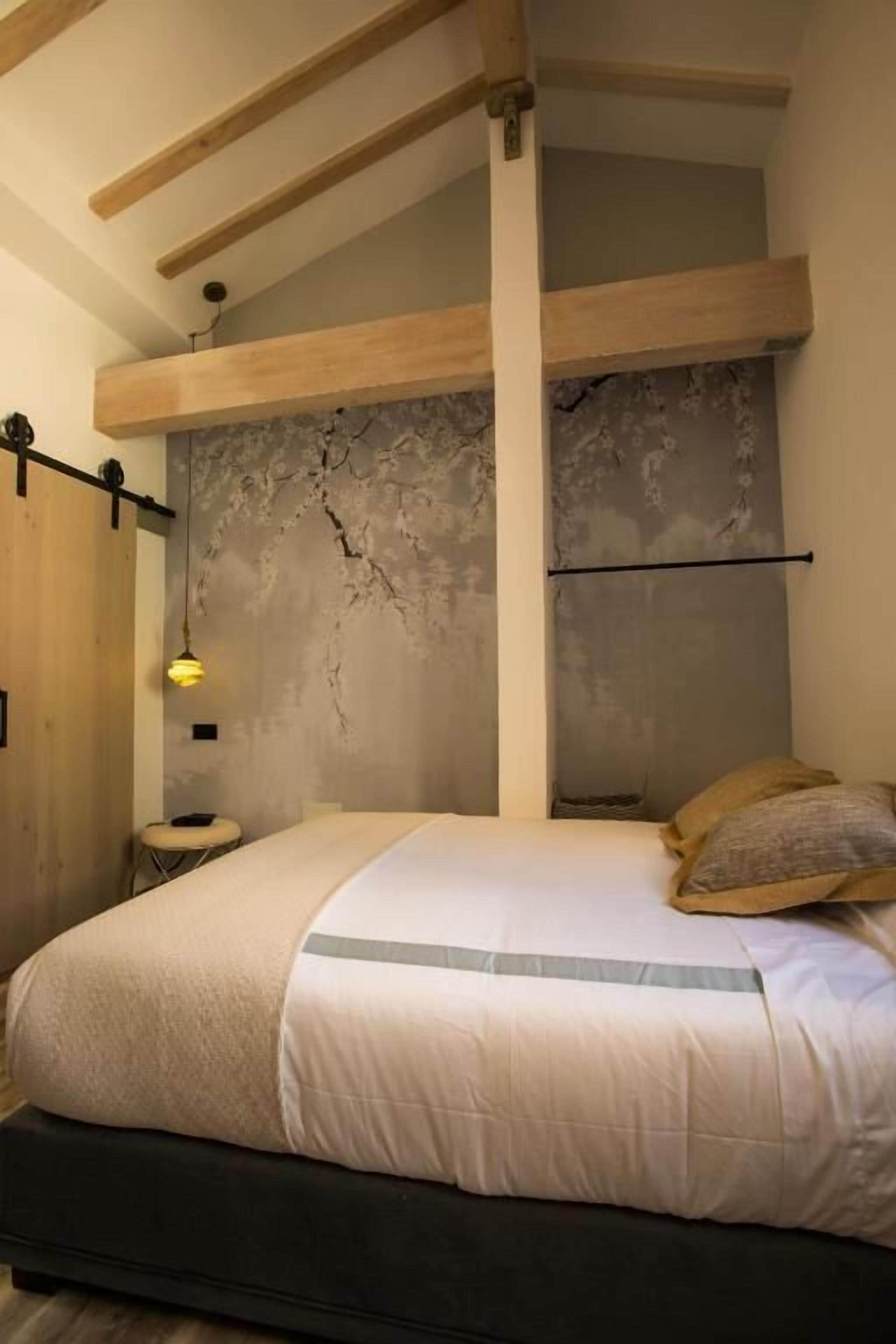 Une chambre à coucher avec un lit, des poutres en bois, un mur en pierre, une petite table et un luminaire suspendu.