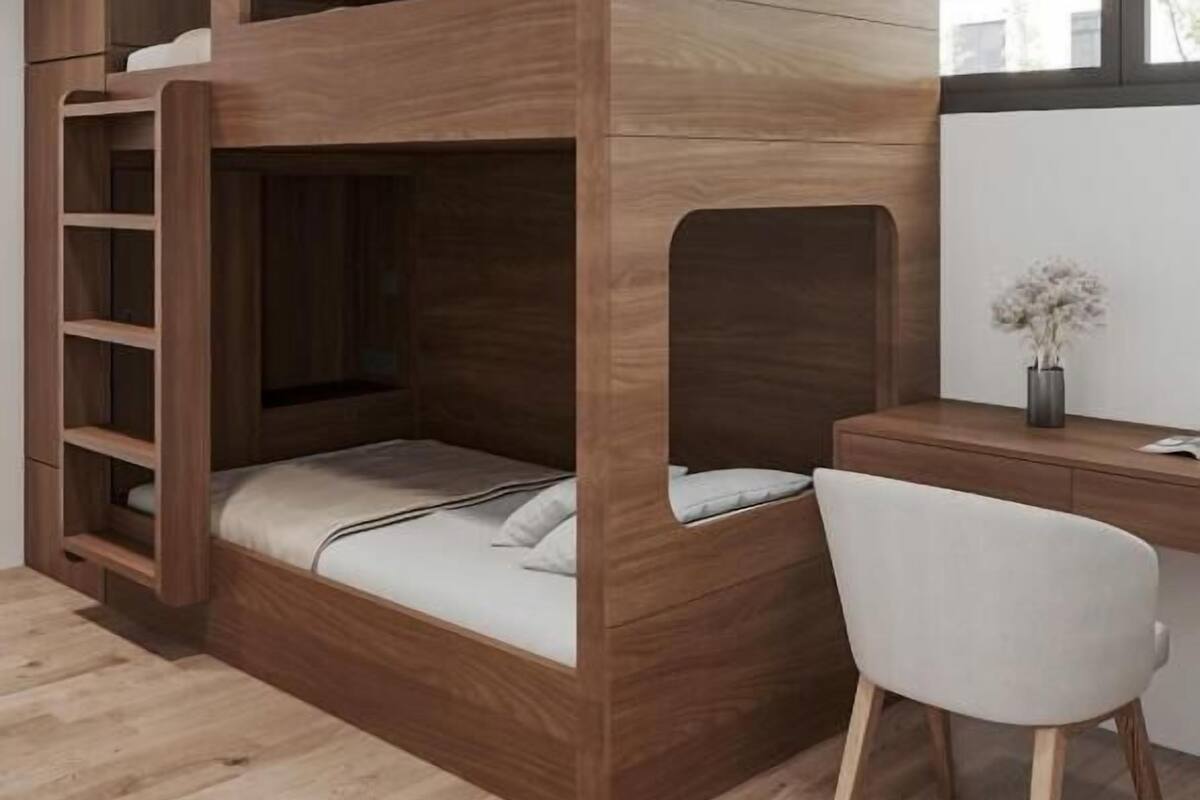A bunk bed with a desk and chair, wooden flooring, and a window with a view of buildings.