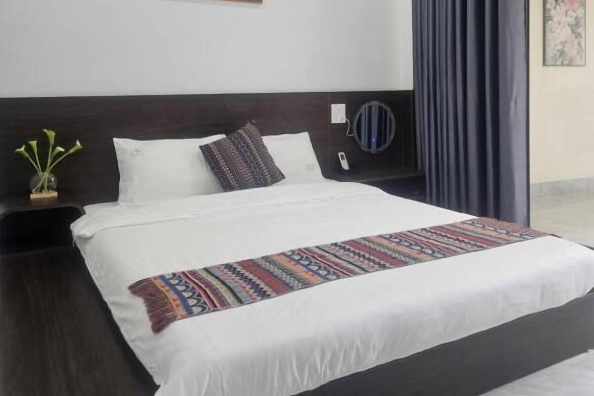 A modern hotel room with a large bed, a painting on the wall, a bedside table with a vase, and a view of the outdoors.