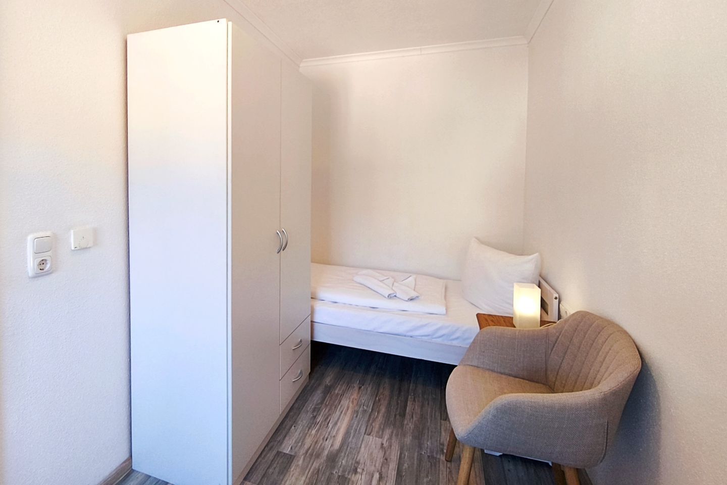 A compact hotel room with a bed, a chair, a wardrobe, and a bedside table with a lamp.