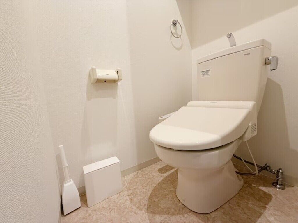 Eine weiße Toilette mit Tank, Toilettenpapierhalter und Bidetanschluss in einem Badezimmer mit beigen Fliesen.