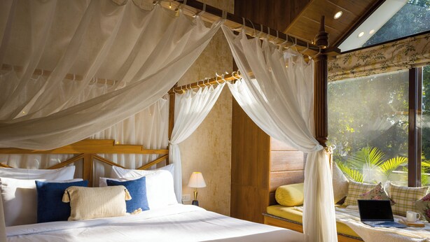 Una cama con dosel, una mesita de noche con un ordenador portátil y una ventana con vistas a zonas verdes.