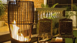 Un patio con pared de ladrillo, una silla de metal junto a un fuego, y dos mesas de madera con copas de vino.