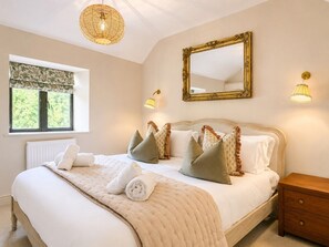 A bedroom with a large bed, a bedside table, a nightstand, a window with a view, and a wall-mounted mirror. - The Potting Shed | Banham Cottages (Bourton-on-the-Water)