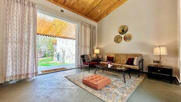 A spacious living room with a wooden ceiling, a large sliding glass door leading to a garden, a sofa set, a coffee table, and a side table with a lamp.