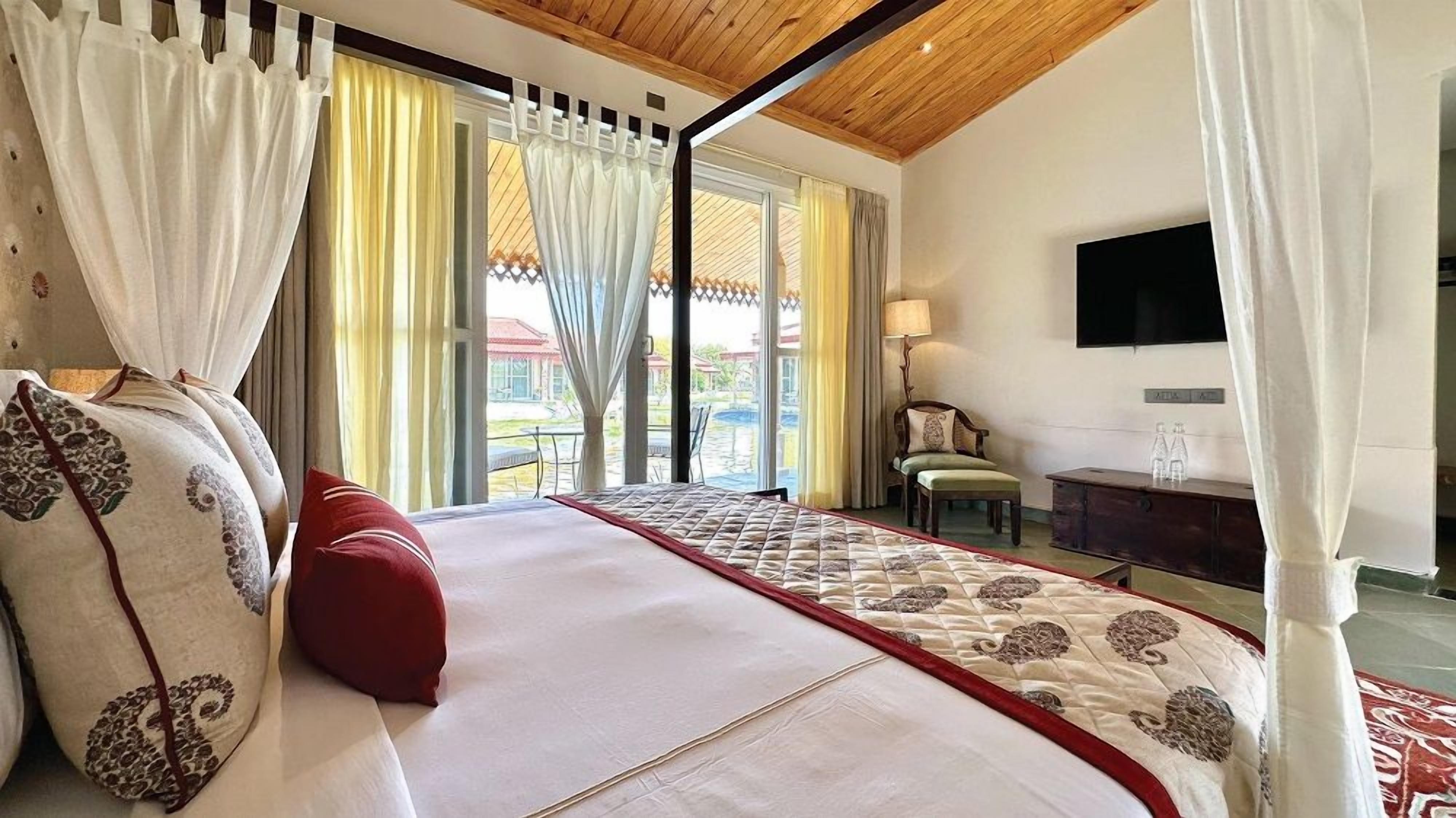 A bedroom with a large bed, curtains, a TV, a desk, and a view of the outside.