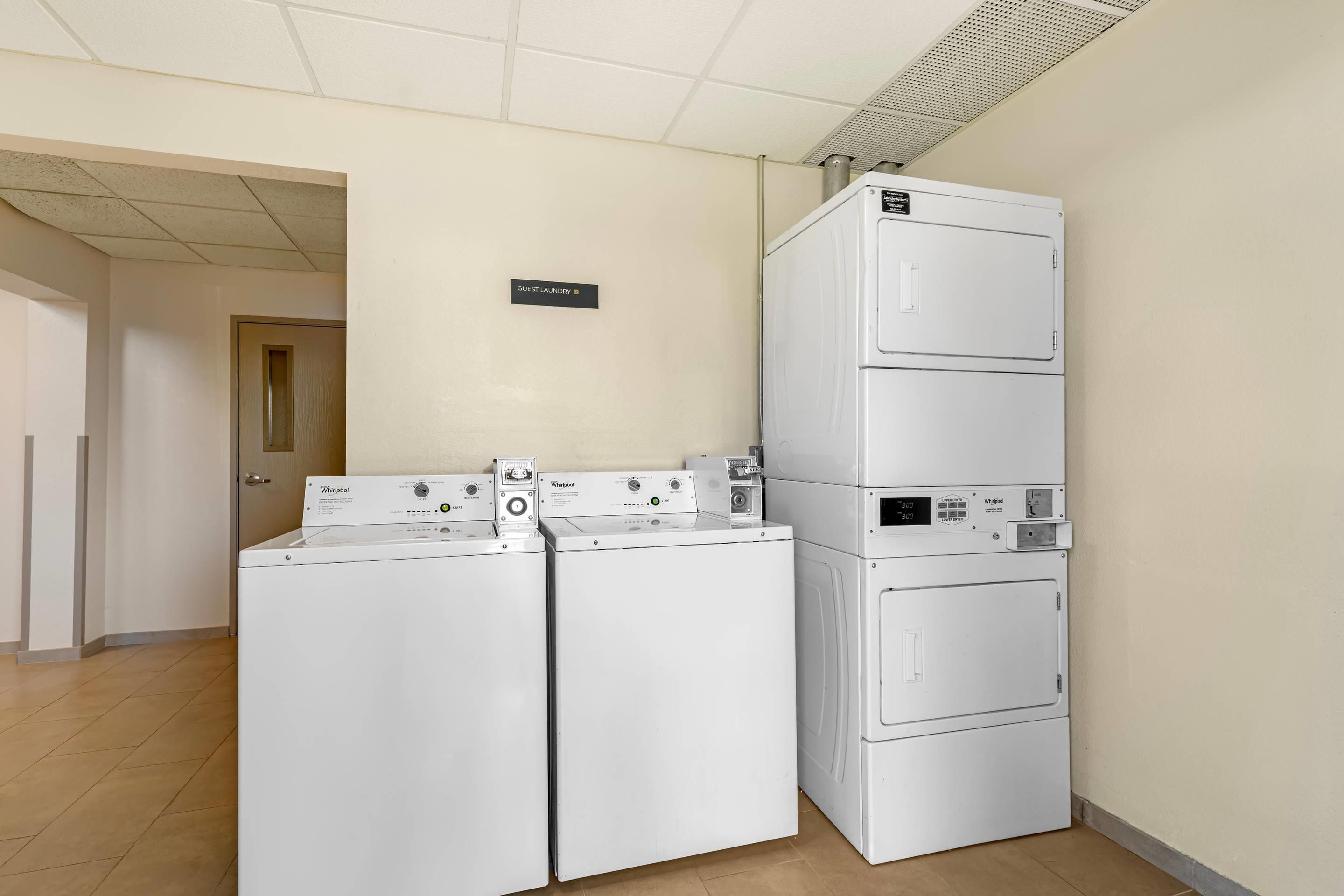 A laundry area with three Whirlpool washers and dryers, a sign indicating guest laundry, and a tiled floor.