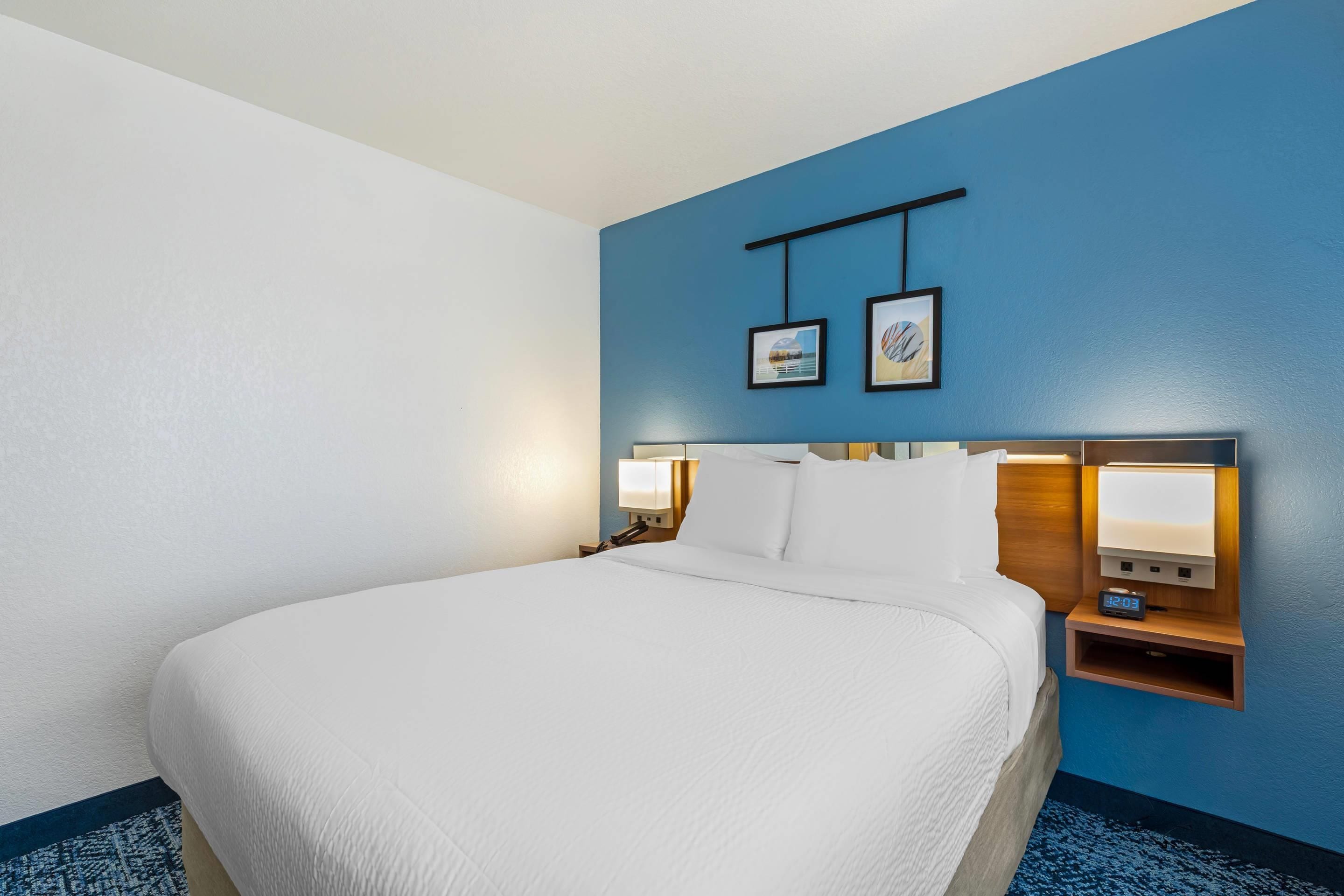 Una habitación de hotel con una cama grande, una mesita de noche con lámpara y dos cuadros en la pared.
