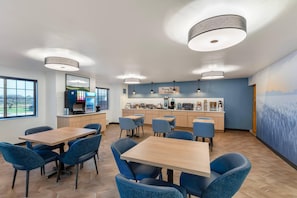 A modern dining area with wooden tables and blue chairs, a kitchen with coffee machines, and a mural on the wall.