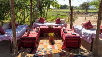 An outdoor dining area with tables set for two, surrounded by trees and a view of a field.