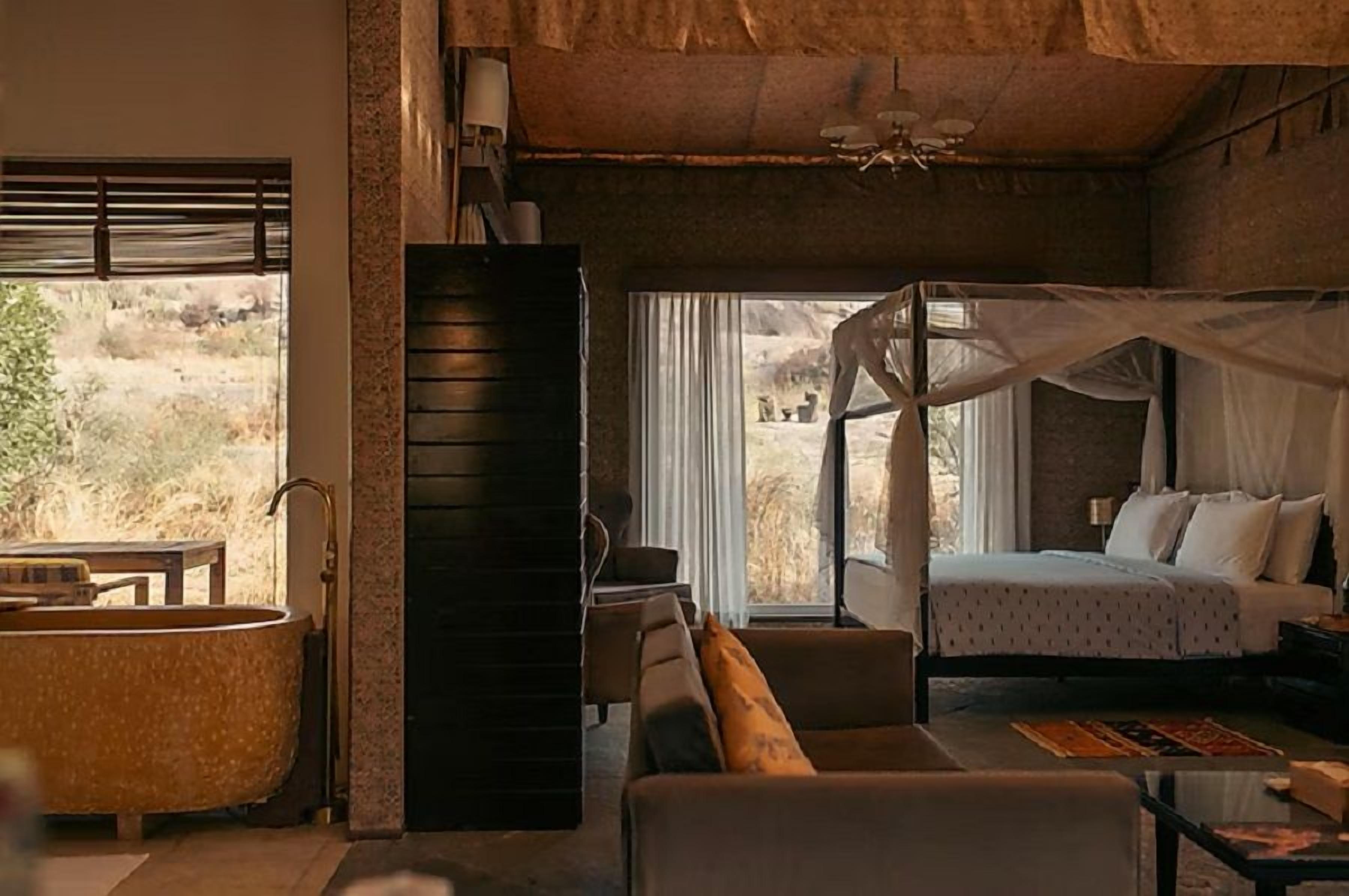 A bedroom with a bed, a nightstand, a bedside table, a large window with curtains, a view of the outdoors, and a bathtub.