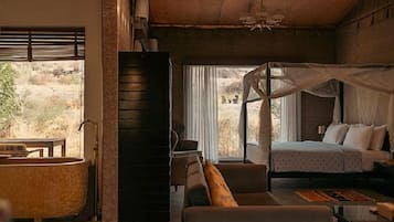 A bedroom with a bed, a nightstand, a bedside table, a large window with curtains, a view of the outdoors, and a bathtub.