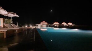 A nighttime swimming pool with lounge chairs and a covered area, surrounded by buildings with red roofs. - Brij Pola,jawai (Bali)