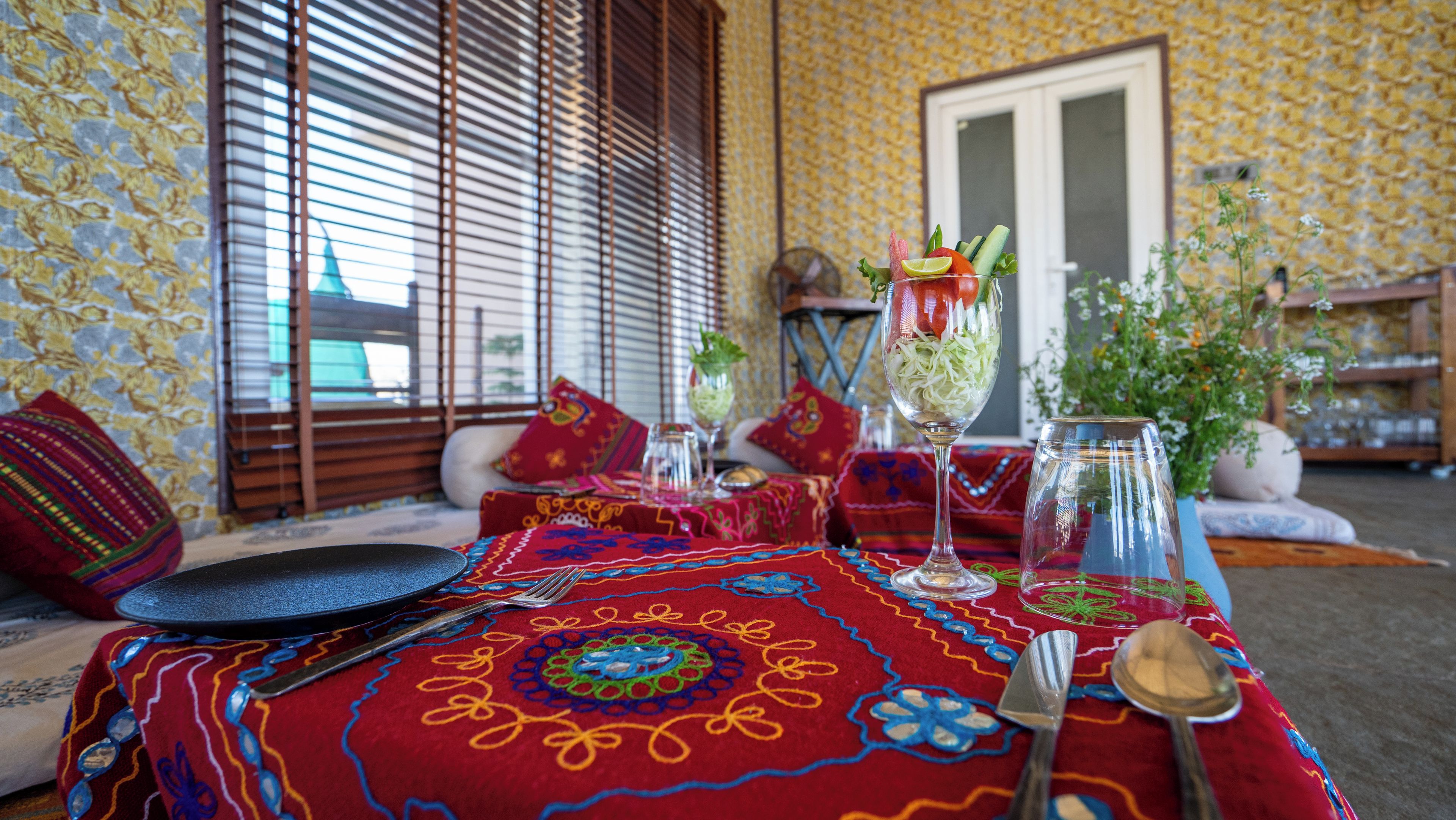 A dining table set with a patterned tablecloth, a glass of water, a glass of juice, and a plate with a spoon.