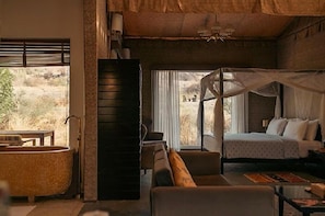 A bedroom with a bed, a nightstand, a bedside table, a large window with curtains, a view of the outdoors, and a bathtub. - Brij Pola,jawai (Bali)