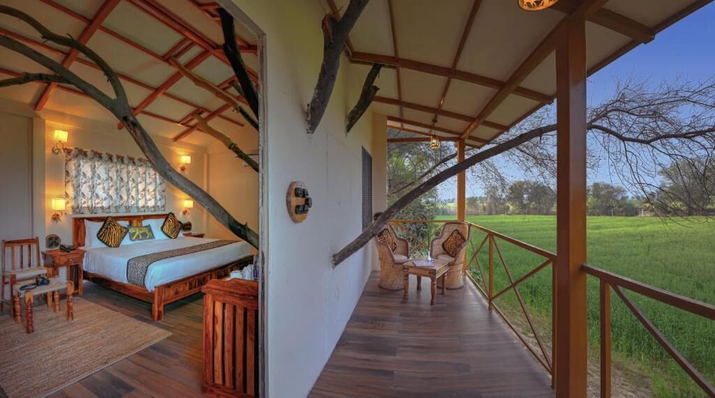 A room with a bed, wooden furniture, a balcony with a railing, and a view of a green field.