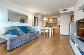 A modern living room with a grey sofa, a dining table, and a kitchen in the background. - Sedna: Apartment with sea views (L'Escala)