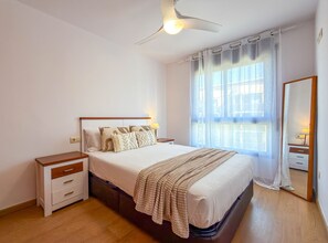A bedroom with a bed, bedside tables, a ceiling fan, a window with curtains, and a mirror. - Sedna: Apartment with sea views (L'Escala)