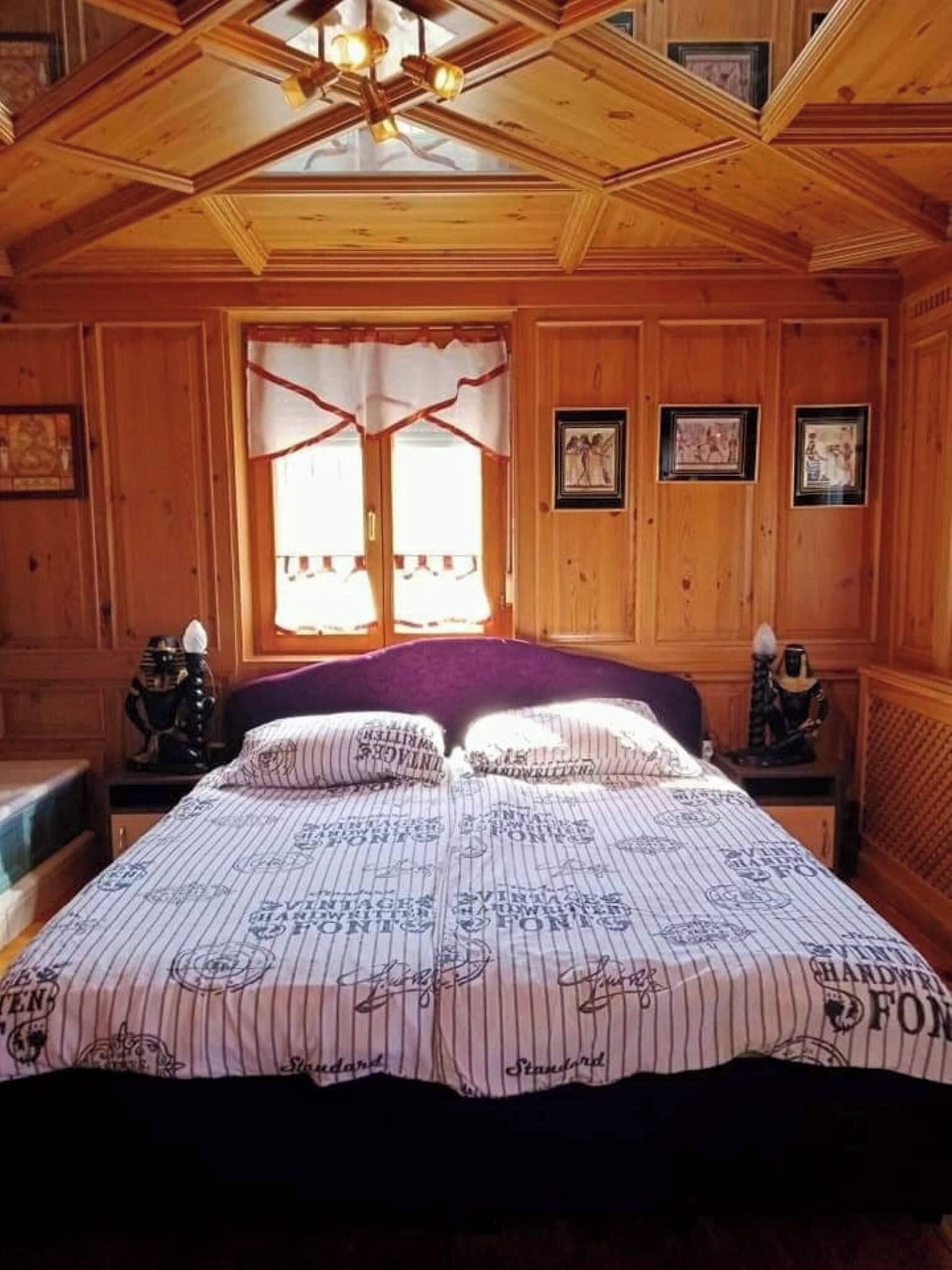 Une pièce aux murs lambrés, avec un lit, une fenêtre ornée de rideaux et des tableaux encadrés.