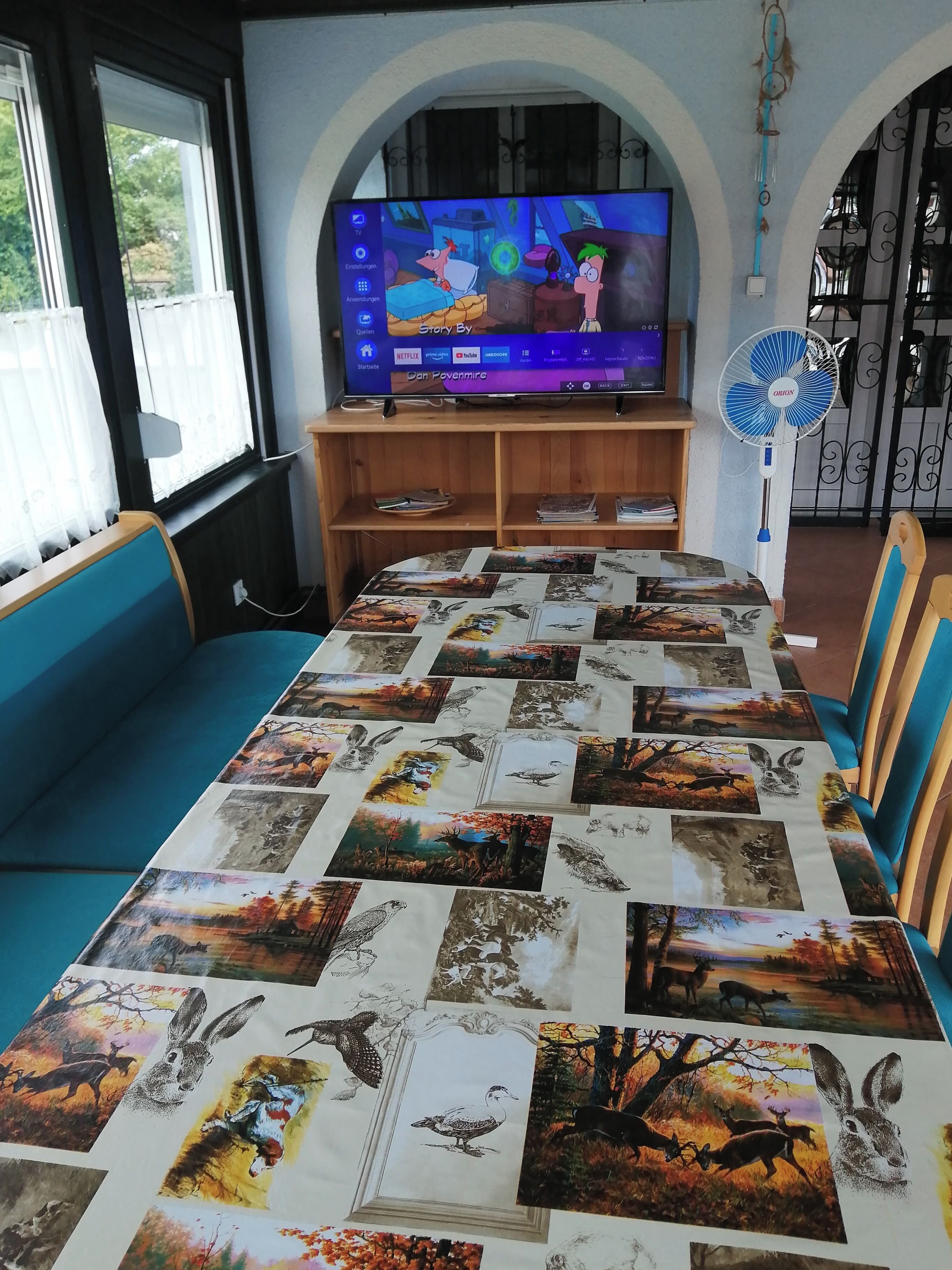 Un salon doté d’un grand téléviseur à écran plat, d’une table recouverte d’une nappe à motifs naturels et d’un ventilateur.