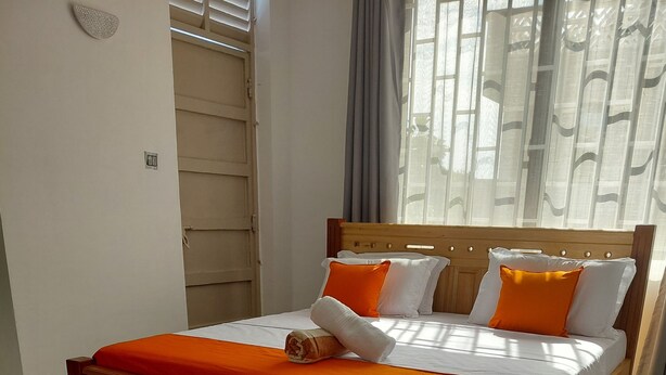 Ein hölzernes Bett mit weißer und orangefarbener Bettdecke, ein Holz-Kopfbrett und ein Fenster mit Vorhängen.