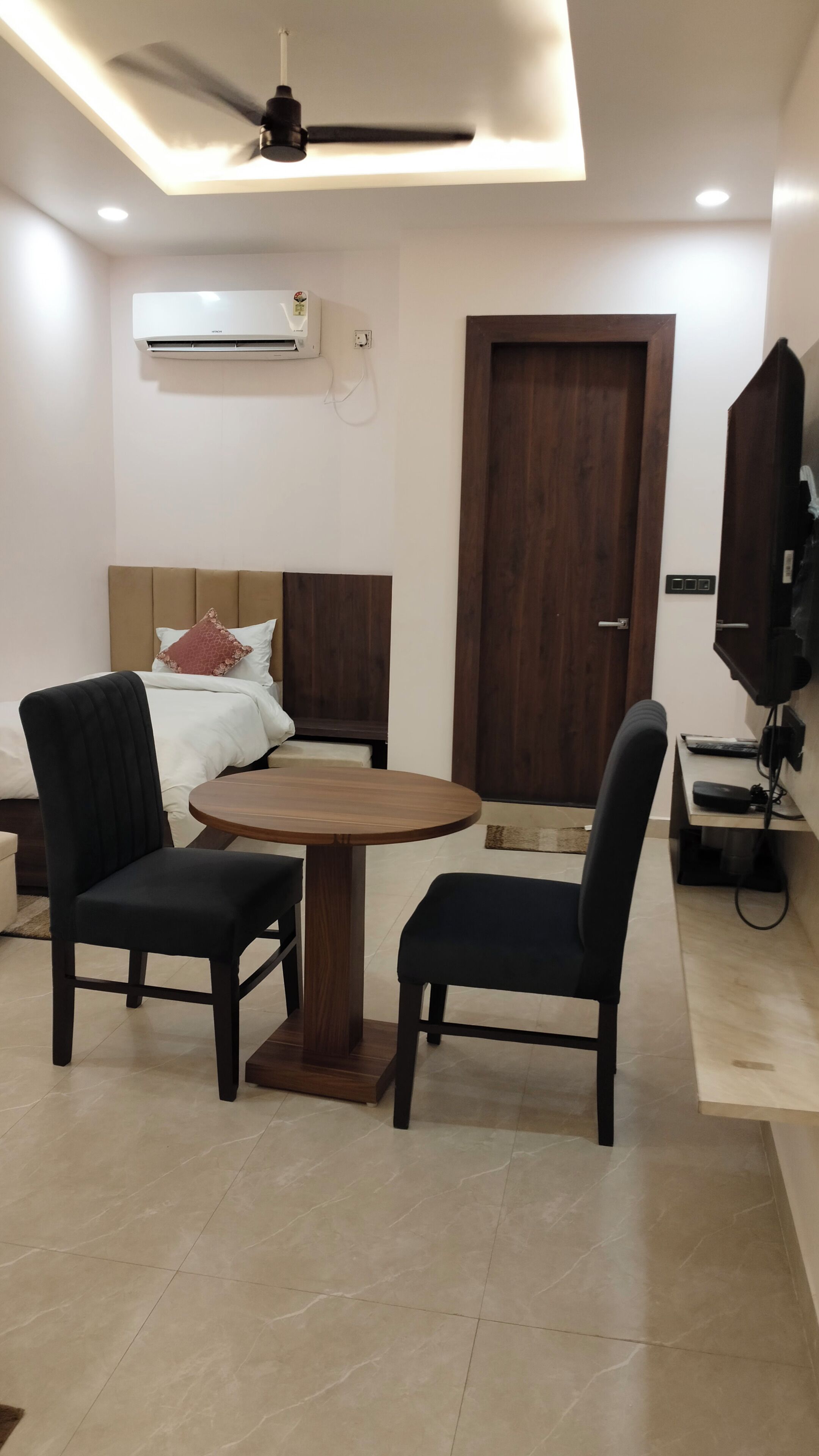A hotel room with a bed, a round wooden table, two chairs, a ceiling fan, an air conditioning unit, a TV, and a door.