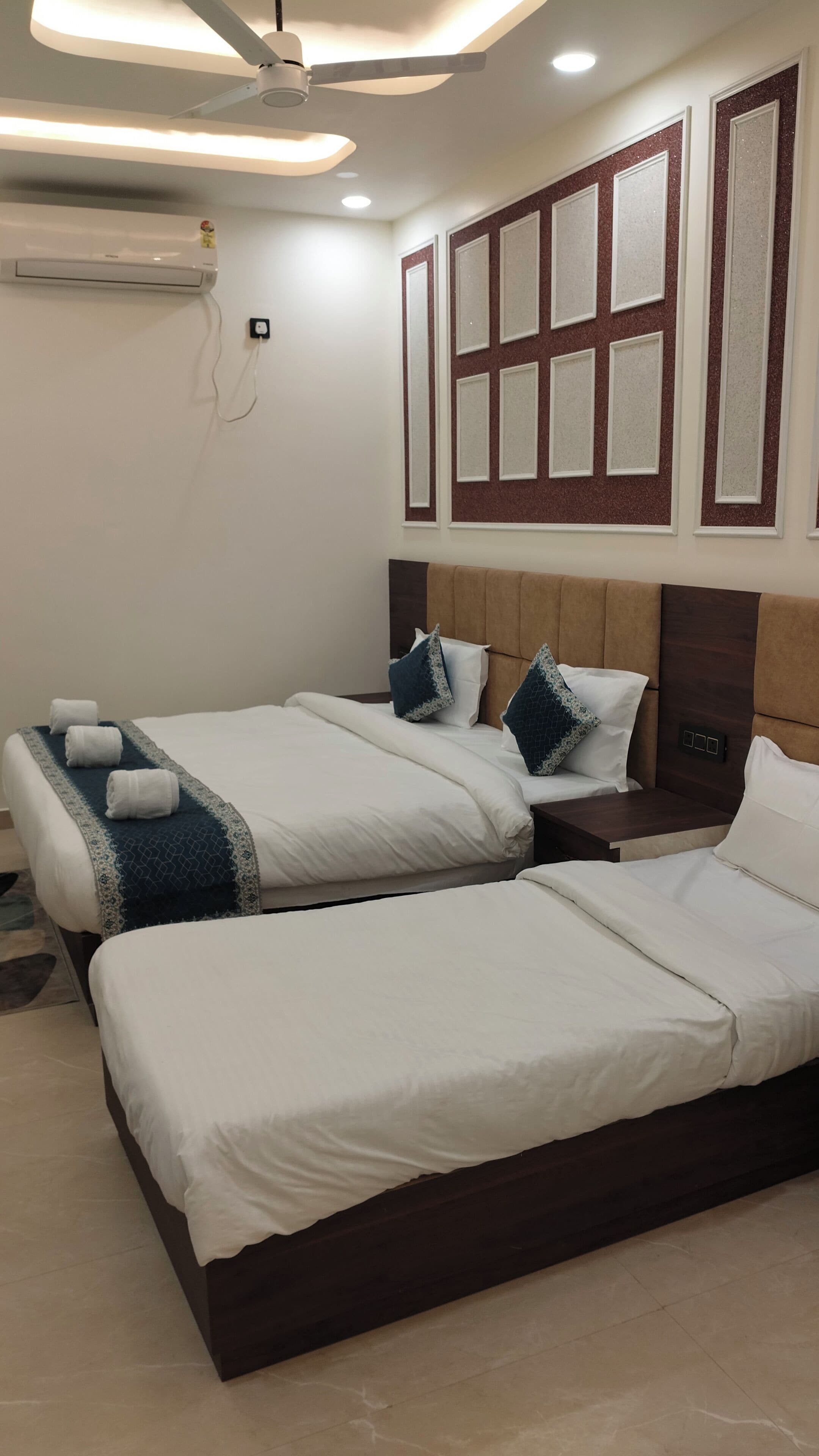 Ein Hotelzimmer mit zwei Betten, einem Deckenventilator, Einbauleuchten, einer Wandklimaanlage und einer Wandverkleidung über den Betten.