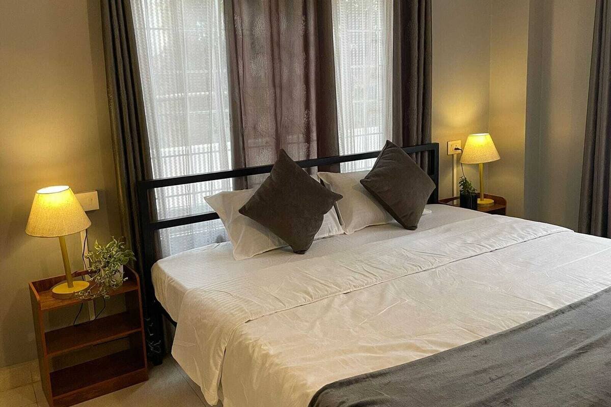 A neatly made bed with white bedding and two pillows, flanked by bedside tables with lamps, in a room with a window and curtains.
