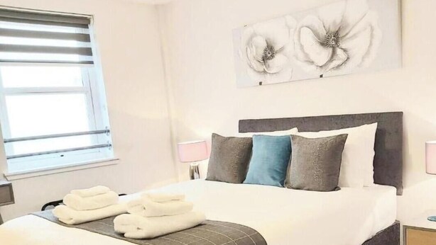 A neatly made bed with a dark headboard, white bedding, and a grey blanket. There are two bedside lamps, a window with blinds, and a wall-mounted artwork of flowers. - Inverness City Centre Apartment With elevator (Inverness)