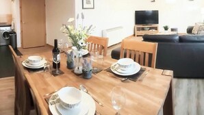A dining area with a wooden table set for four, a vase of flowers, and a television in the background. - Inverness City Centre Apartment With elevator (Inverness)