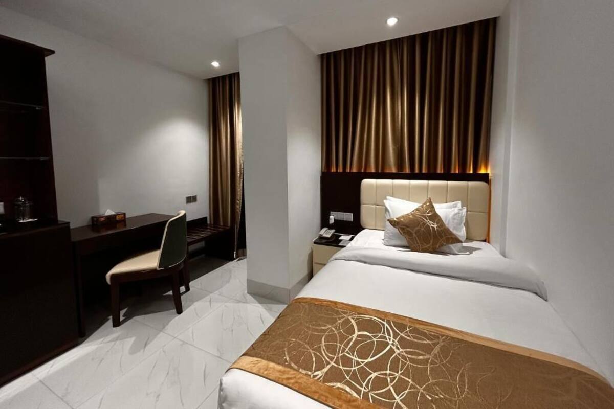 A hotel room with a bed, a desk, a chair, a nightstand, and a wall with vertical blinds.