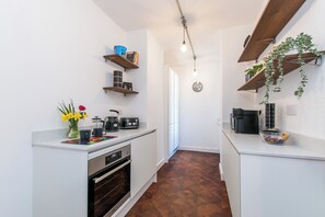 Cozinha compacta com armários brancos, forno embutido, cafeteira e relógio na parede.