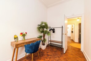 Quarto com escrivaninha de madeira, uma cadeira azul, planta, cabideiro e porta para outro cômodo.