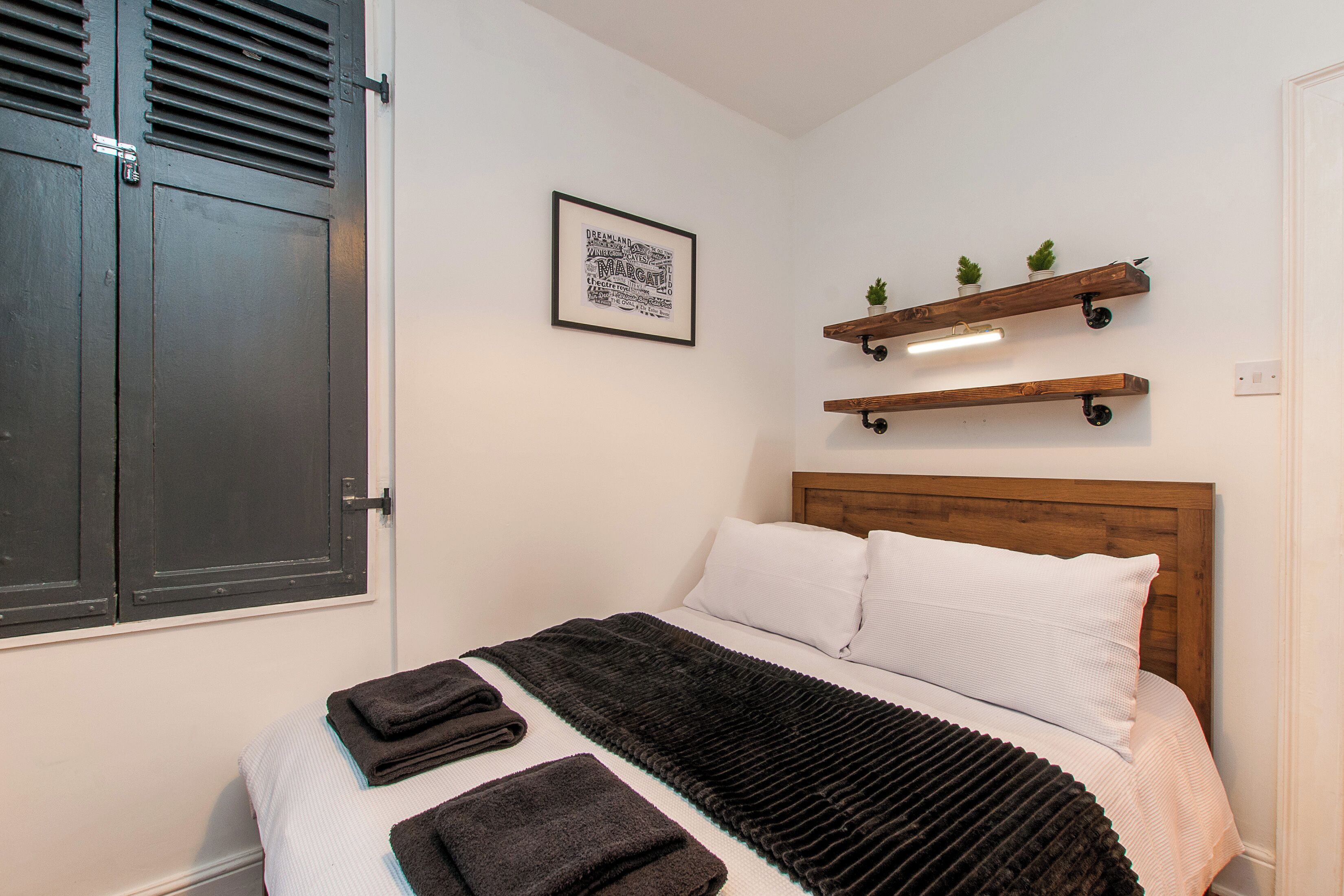 A bedroom with a bed, a headboard, a wall-mounted shelf with plants, a framed picture, and a door with a handle.
