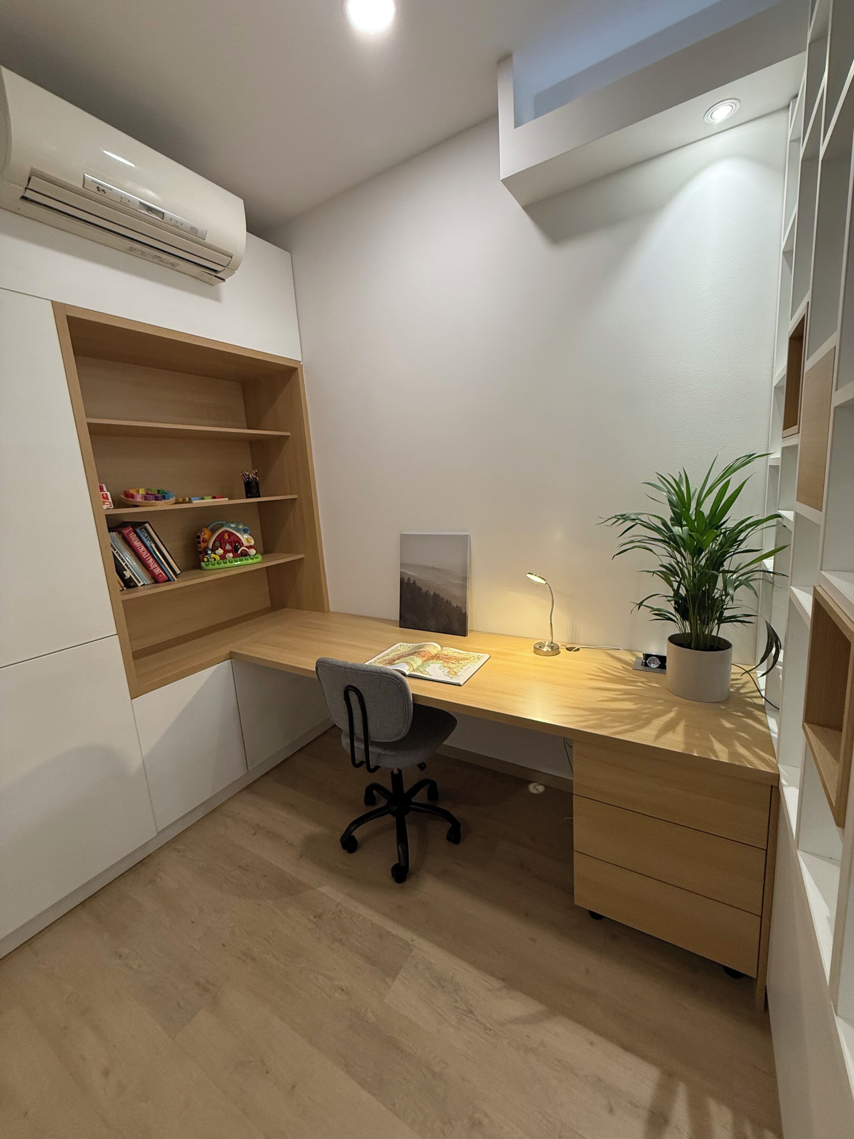 A modern study room with a wooden desk, a black office chair, a bookshelf, a potted plant, and a ceiling-mounted air conditioner.