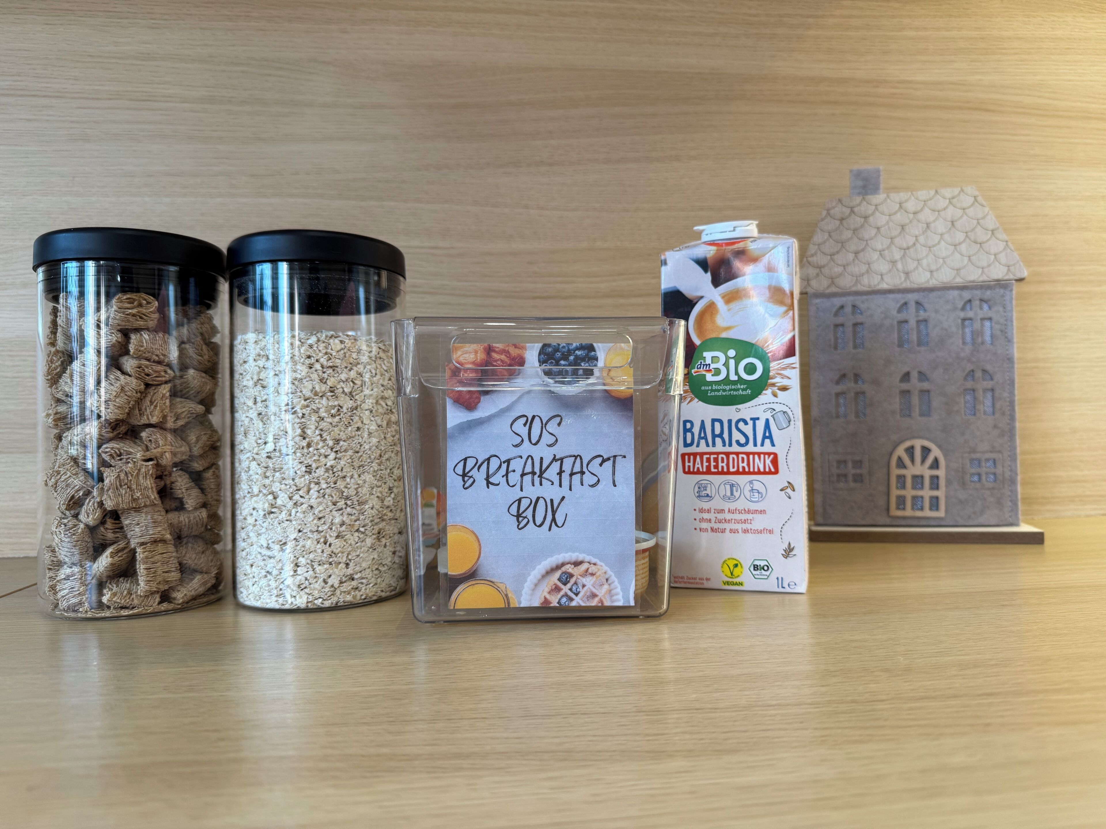A breakfast box, two containers of grains, a carton of Barista Haferdrink, and a model house.