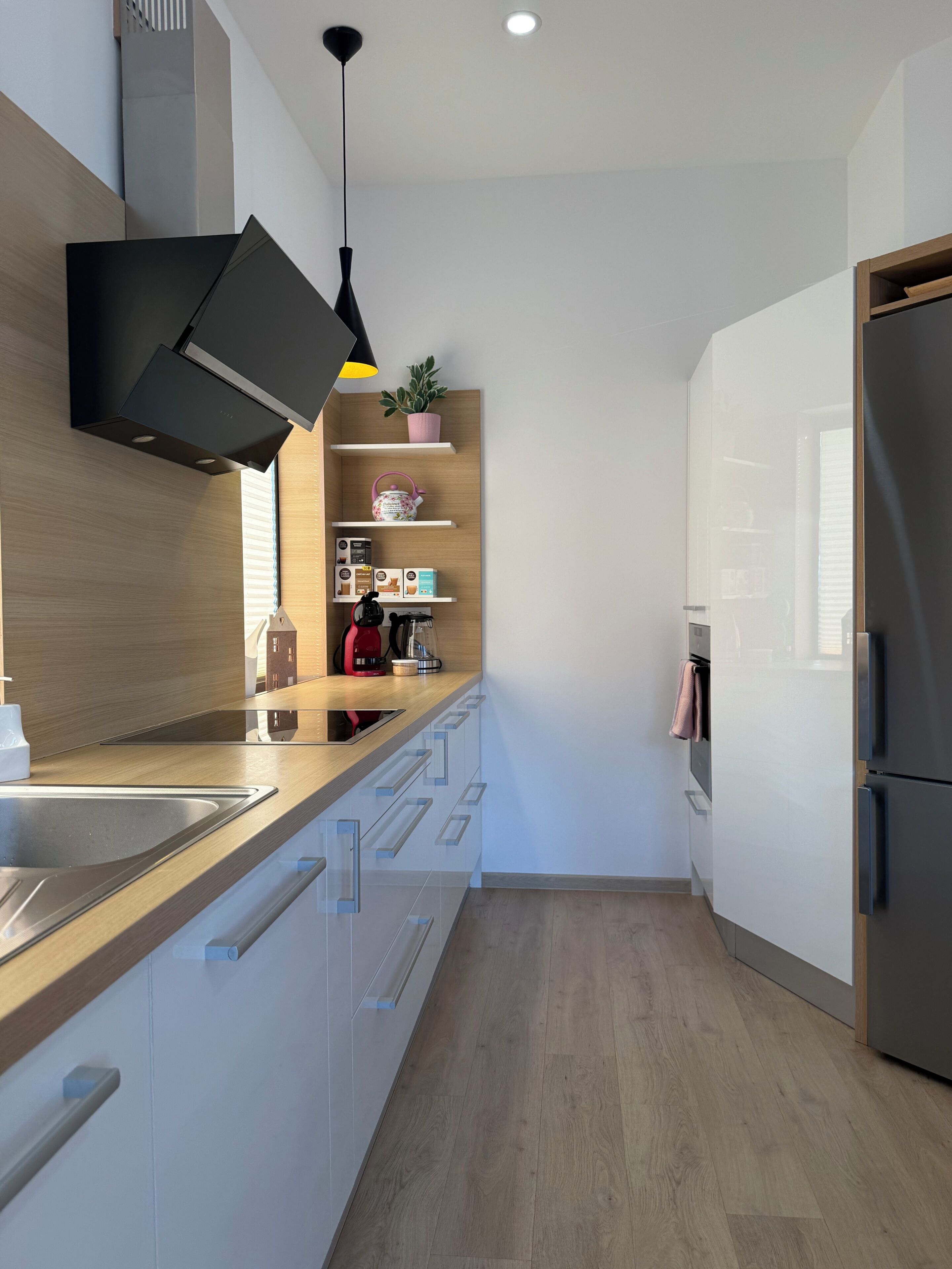 A modern kitchen with a built-in oven, a stainless steel sink, wooden cabinets, and a black pendant light.