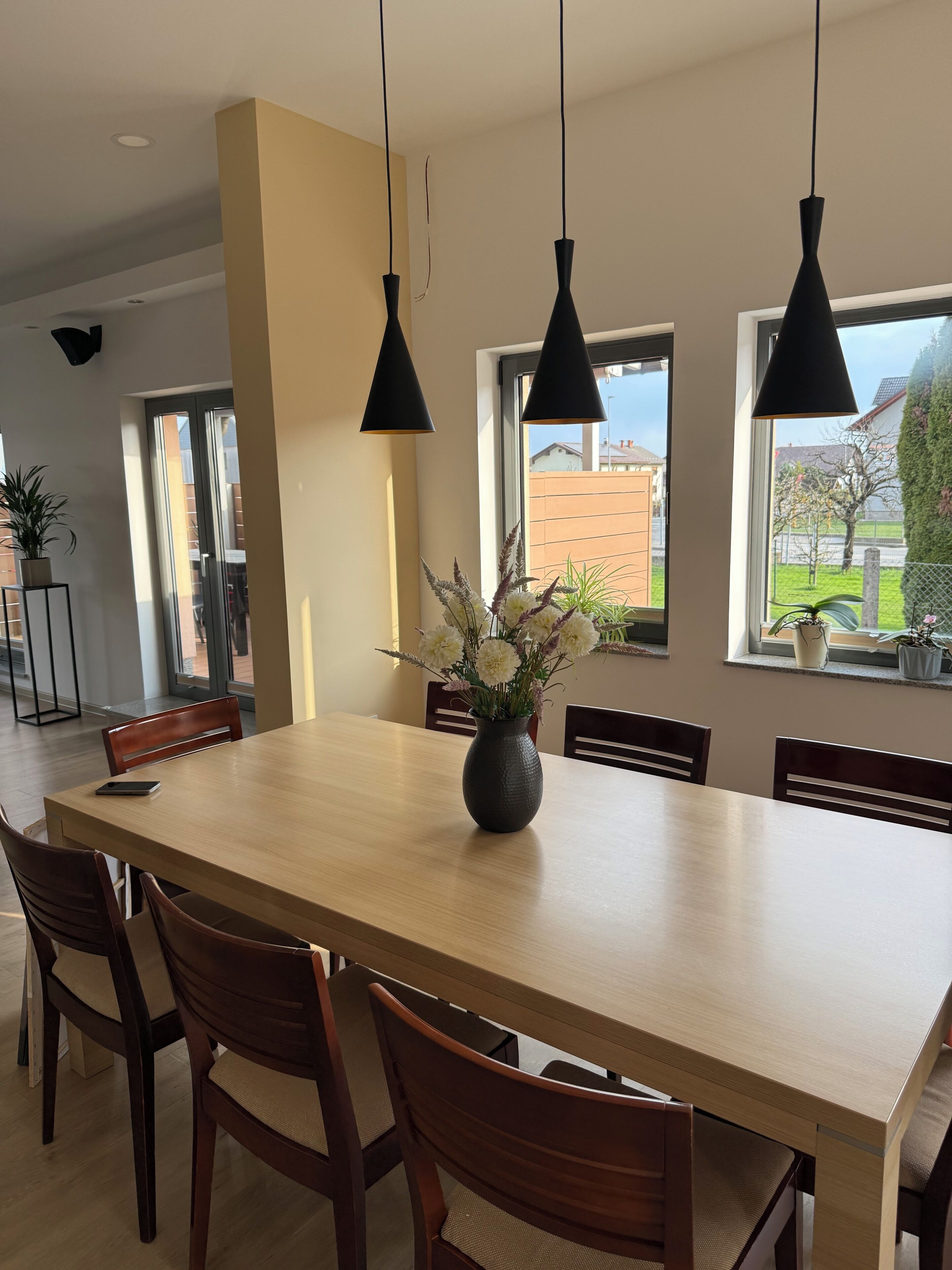 Un comedor con una mesa larga, sillas, lámparas colgantes, un jarrón con flores y ventanas con vistas al exterior.