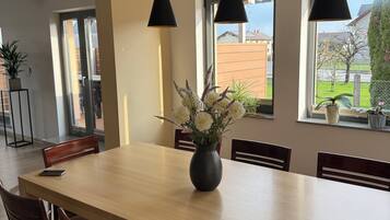 Un comedor con una mesa larga, sillas, lámparas colgantes, un jarrón con flores y ventanas con vistas al exterior.