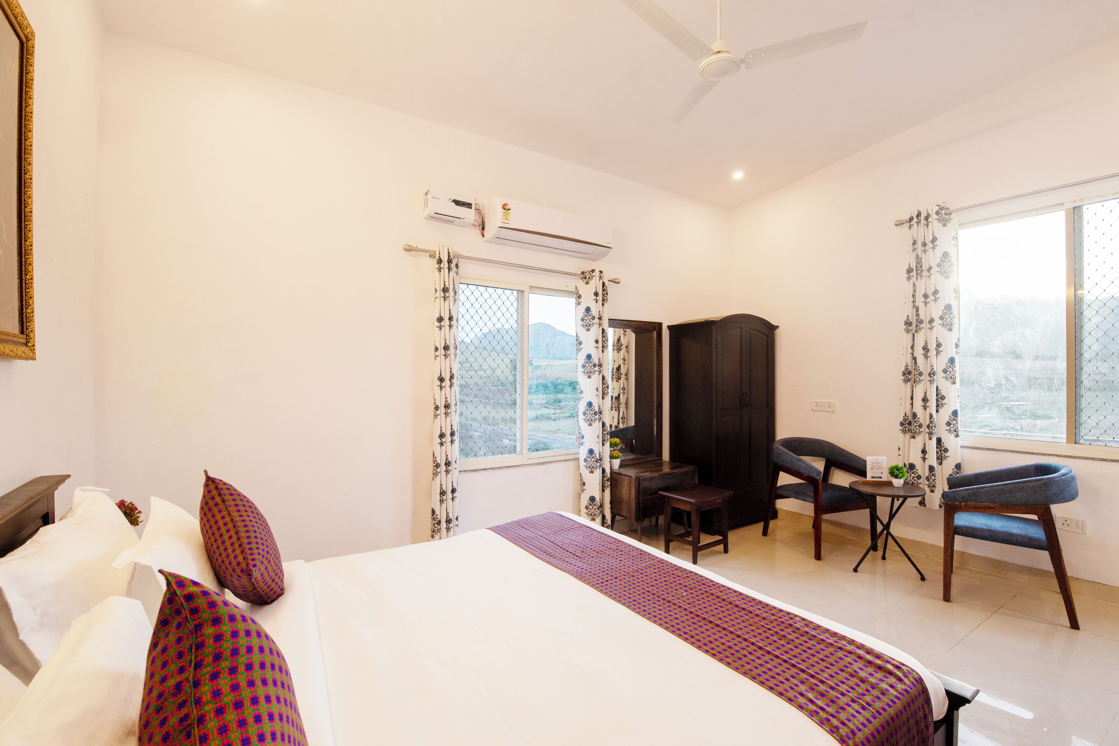 A hotel room with a bed, a desk, two chairs, an air conditioning unit, and a window with curtains.